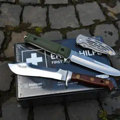 Drei Messerarten, von Jagdmessern bis zu Klappmessern, liegen auf einer Erste-Hilfe-Box. Diese Messer sind vielseitige Werkzeuge und können auch als Waffen dienen.