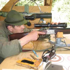 Ein Mann mit Hut zielt mit einem Gewehr mit kurzem Lauf. Auf dem Tisch liegen ein Tacker und Zielscheiben.