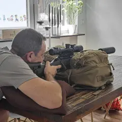 Mann im jagdlichen Anschlag mit Gewehr und Zielfernrohr auf einem Tisch.