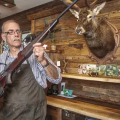 Ein Mann prüft eine Blaser R8 Büchse, vielleicht wegen Blaser R8 Verschluss Probleme. Ein Hirschkopf hängt an der Wand.