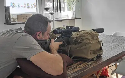 Mann im jagdlichen Anschlag mit Gewehr und Zielfernrohr auf einem Tisch.