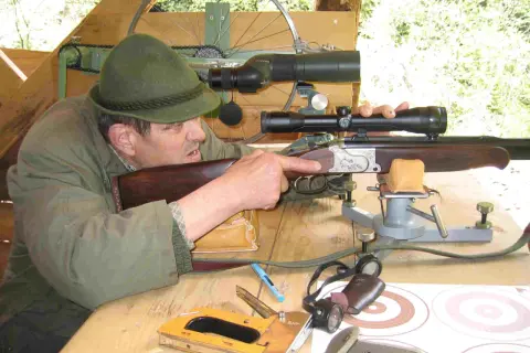Ein Mann mit Hut zielt mit einem Gewehr mit kurzem Lauf. Auf dem Tisch liegen ein Tacker und Zielscheiben.