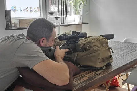 Mann im jagdlichen Anschlag mit Gewehr und Zielfernrohr auf einem Tisch.