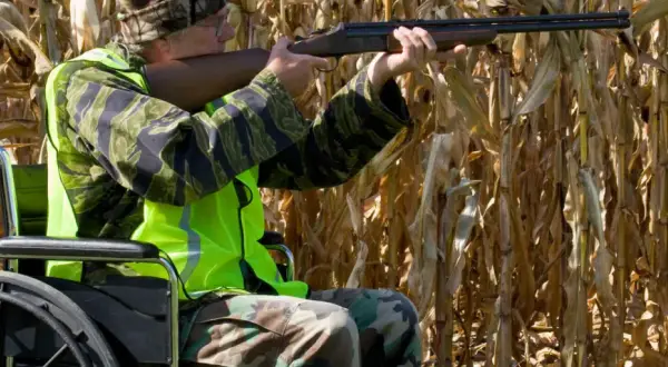Jäger im Rollstuhl mit Gewehr im Maisfeld, bekleidet mit Tarnung und Warnweste. Dies könnte unter paragraph 6 waffengesetz fallen.