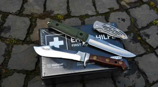 Drei Messerarten, von Jagdmessern bis zu Klappmessern, liegen auf einer Erste-Hilfe-Box. Diese Messer sind vielseitige Werkzeuge und können auch als Waffen dienen.