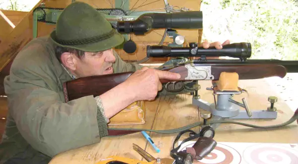 Ein Mann mit Hut zielt mit einem Gewehr mit kurzem Lauf. Auf dem Tisch liegen ein Tacker und Zielscheiben.