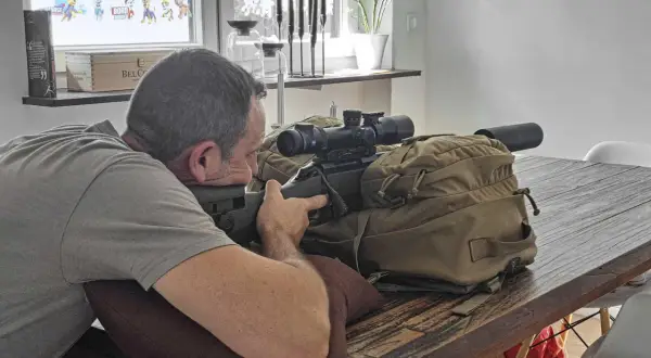 Mann im jagdlichen Anschlag mit Gewehr und Zielfernrohr auf einem Tisch.