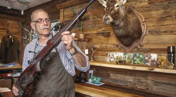 Ein Mann prüft eine Blaser R8 Büchse, vielleicht wegen Blaser R8 Verschluss Probleme. Ein Hirschkopf hängt an der Wand.
