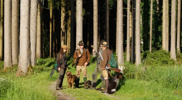 Vier Jäger mit Hunden im Wald. Einer wirkt wie ein jagdgast ohne begehungsschein, da er nur mit Fernglas und ohne Gewehr zu sehen ist.