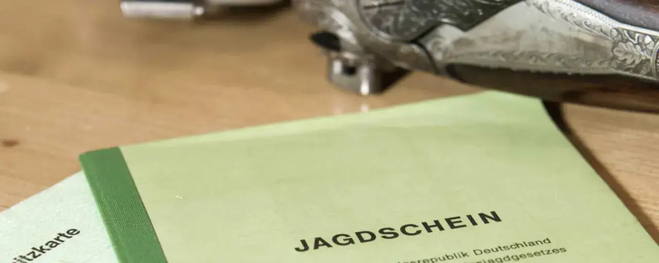 Jagdschein und eine Schusswaffe liegen auf einem Holztisch. Dies illustriert die Anforderungen des § 6 Waffengesetz für Jäger.