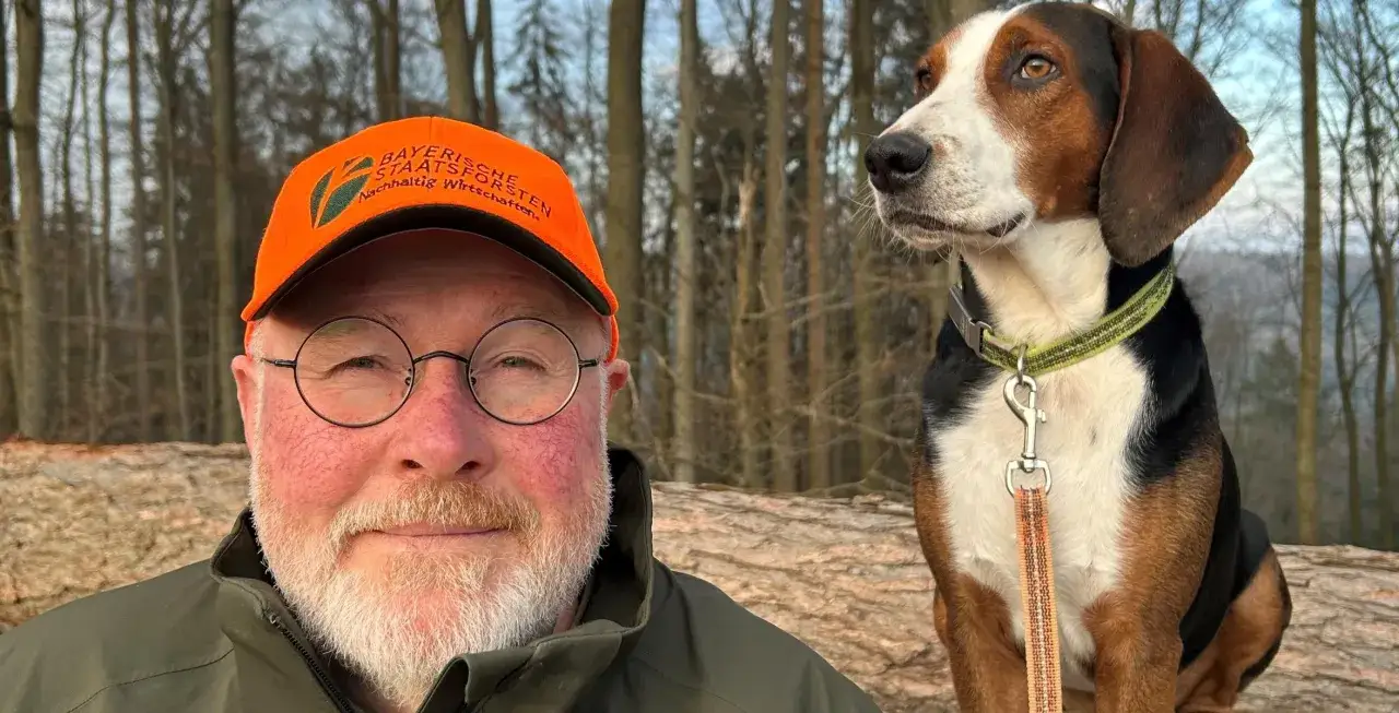 Ein Mann mit Brille und orangefarbener Kappe sitzt neben einem treuen Waidslaut Hund im Wald.